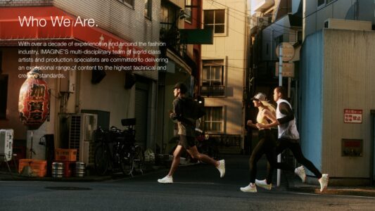 Three people are jogging down an urban street flanked by buildings in warm, late afternoon light. Their athletic wear contrasts with the surrounding environment, which includes bicycles, crates, and a traditional lantern. Shadows are cast from nearby structures, adding depth to the scene. The atmosphere feels dynamic and vibrant, emphasizing movement and the merging of urban life with fitness.