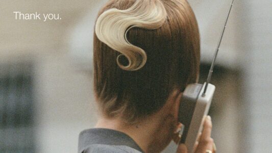 A person is holding a vintage mobile phone with an extended antenna to their ear. Their back is facing the viewer. The person has short brown hair with a distinctive curled blonde streak. The attire is a gray outfit, and the setting appears to be an urban environment, slightly blurred in the background for depth.