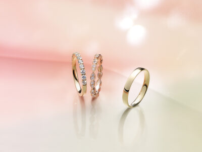 Three elegant rings are displayed against a softly blurred pink and beige background. The left ring features a row of sparkling gemstones. The center ring has a twisted design, adding a unique touch. The right ring is a simple, classic gold band. The arrangement exudes luxury and sophistication, with light reflections enhancing the gleaming surfaces.