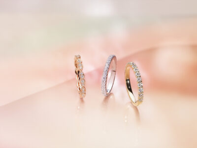 Three elegant rings are artfully displayed on a soft, blurred background in shades of peach and cream. The first ring features a continuous row of round gemstones set in a rose-gold band. The second ring is encrusted with sparkling gems on a white-gold band. The third ring combines a classic yellow-gold band with a row of shimmering stones.