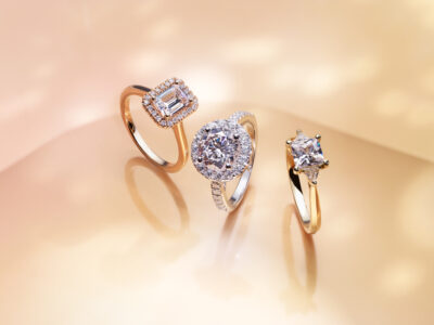 Three elegant rings are showcased on a soft, glowing background, each featuring a prominent gemstone. The first ring displays a rectangular gem surrounded by smaller stones, set in a gold band. The second ring highlights a round gem with a halo of smaller stones. The third has a square gem with supporting prongs, all exuding luxury and sophistication.