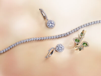 Sparkling elegance defines this arrangement, showcasing a delicate bracelet, a pair of earrings, and a ring. The bracelet is adorned with a continuous strand of shimmering stones. The earrings and ring feature large, brilliant gems encircled by smaller stones, with the ring incorporating vibrant green elements. The soft, blurred background enhances the luxurious, radiant quality of the pieces.