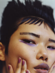 A person with short, spiky dark hair and bold makeup is portrayed. The eyes are accentuated with purple eyeshadow, and the eyebrows are defined. Their lips are shaded in a similar hue. Their fingers, with matching purple nail polish, gently touch their cheek, creating a sense of introspection and style. The overall tone suggests a modern, edgy aesthetic.