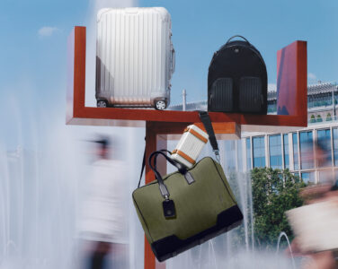 A silver suitcase, black backpack, and olive duffle bag are artfully arranged on a wooden structure. The backdrop features a clear sky and modern architecture, with blurred figures suggesting movement. Water from nearby fountains creates a dynamic, outdoor atmosphere. The scene blends fashion and function, highlighting travel essentials against an urban landscape.