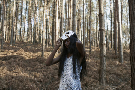 A person stands in a densely wooded area, surrounded by tall, straight trees with slender trunks. The ground is covered in brown foliage. They have long, dark hair and are wearing a patterned outfit, adjusting a cap on their head. Sunlight filters through the trees, creating a serene and natural atmosphere in the forest setting.