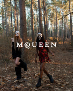 Two individuals wearing masks walk confidently through a dense forest. The first person sports a black outfit with high boots, while the second wears a red plaid dress paired with black boots. Tall trees surround them, and the ground is covered in fallen leaves, capturing an autumnal atmosphere. The scene suggests mystery and adventure in a natural setting.