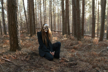 A figure with long hair and a mask kneels on the forest floor, surrounded by tall, slender trees in a woodland setting. The ground is covered with dry leaves and ferns, and sunlight filters through the trees, casting soft light and shadows. The scene appears both mysterious and serene, evoking a sense of solitude and nature.