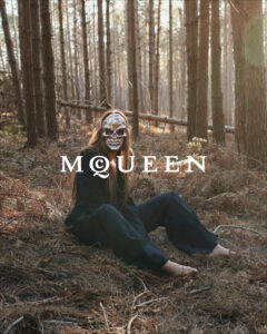 Amidst a dense forest, a figure is seated on the ground covered in dry leaves and branches. Wearing a dark outfit, they don an intricate metallic skull mask. Sunlight filters through the tall, slender trees, casting a soft glow and creating an eerie yet serene atmosphere. The overall scene evokes a sense of mystery and intrigue.
