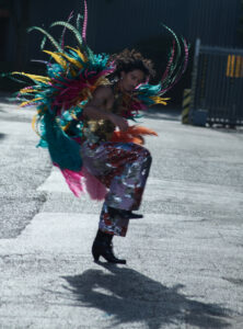 A man dressed in a vibrant, feathered costume bursts with color, incorporating turquoise, yellow, pink, and purple hues. He strikes a dynamic pose mid-motion, exuding energy. His attire includes shiny, multicolored pants and black boots, while the background features an industrial setting with concrete and metal elements, casting distinct shadows on the ground.