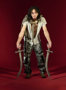 A person with curly hair stands confidently against a vibrant red backdrop, holding two ornate swords. They wear a short-sleeved shirt, patterned trousers, and brown shoes. A draped scarf adds a layer of texture and style. The overall composition conveys a sense of drama and strength, blending elements of fashion with a hint of theatrical flair.
