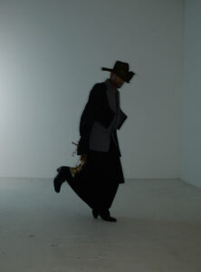 A person dressed in a dark, stylish outfit with a wide-brimmed hat and a textured blazer holds a shiny, golden French horn while stepping forward in an empty, minimalistic room. The composition creates a dramatic and mysterious atmosphere, emphasizing the individual's unique fashion sense and suggestive movement amidst the stark background.