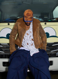 A person with a rugged brown jacket and a multicolored cap is sitting on the front of a vehicle, head bowed. They wear a patterned, torn white shirt underneath the jacket. Below, oversized blue pants with red stitching are visible. The vehicle behind is adorned with large painted eyes, creating a playful, animated facade.