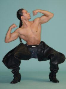 A muscular man with long hair poses confidently, flexing his biceps and looking to the side. He's shirtless, wearing oversized black leather pants and matching boots. The background is a solid teal color, creating a striking contrast with his attire. His dynamic stance emphasizes strength and style, blending elements of fashion and athleticism in an eye-catching presentation.