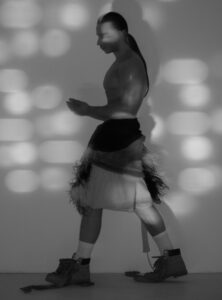 A person with long hair is captured in a dynamic pose, casting a distinct shadow on a textured wall. They wear a unique outfit featuring layered fabric with feather-like details and sturdy boots, creating an intriguing contrast. Soft, patterned light adds depth, playing across the scene and highlighting the person's form against the plain background.