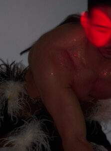 A person is depicted with a partially visible face bathed in red light, adding a mysterious and dramatic effect. The bare shoulder and muscular arm are sprinkled with glitter, catching reflections in the soft lighting. Feathers with varied textures adorn the outfit, contributing to an ethereal, theatrical atmosphere. The composition emphasizes contrast and mood.