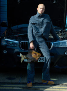 A person with a bald head stands in front of a car with an open hood, dressed in a fashionable, blue patterned two-piece outfit. They hold a unique, fish-shaped bag in one hand. The scene is set outdoors, accentuated by the natural lighting that highlights the details of the clothing and accessories.