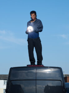 A person stands on top of a black vehicle, against a clear blue sky. Dressed in dark clothing, they hold a glowing object, which casts a bright light. Their posture is relaxed yet focused, directing attention toward the spherical luminescence. The scene evokes a sense of curiosity, blending ordinary surroundings with an element of mystery.