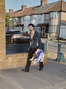 A stylish individual walks along a suburban street, dressed in a partially open, striped shirt and dark trousers. They hold a bundle of colorful fabric and objects. The background features a row of residential houses with bay windows, red-tiled roofs, and parked cars, while a clear blue sky enhances the serene neighborhood atmosphere.