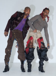 Two individuals pose in avant-garde fashion, exuding an artistic and eccentric vibe. One sports a leather jacket with bold patterns over a purple top, pairing it with brown trousers and lace-up boots. The other wears a striped sweater with patched pants, red details, and high black boots. Both hold phones, showcasing distinct personal styles and expressions.