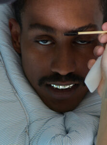 A person with a focused expression is wrapped in a blue and white striped fabric. They wear shiny metallic dental grills, enhancing a confident look. A hand holds a spoolie brush near their eyebrow, suggesting grooming or styling. The overall atmosphere is intimate and detailed, highlighting personal care and bold accessorizing choices.