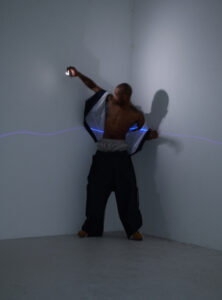A shirtless man stands in a minimalist corner space, wearing oversized pants and tan shoes. His jacket, partially open, reveals a streak of blue light across his chest. His arm is outstretched, holding a small light source that casts shadows on the wall, adding a dynamic and mysterious atmosphere to the scene.