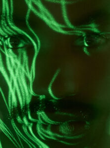 Shimmering green light patterns intricately dance across a face, highlighting features such as eyes and a mustache. The abstract illumination creates a mysterious, almost digital aesthetic. The interplay of shadow and light casts an ethereal glow, emphasizing contours and expressions, resulting in a captivating, surreal portrait that blends organic and futuristic elements in a harmonious display.