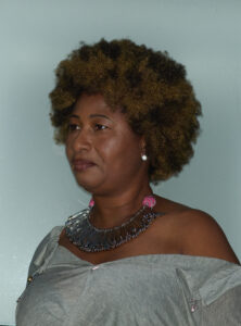 A person with curly hair is facing forward, wearing a gray off-the-shoulder garment. They have a confident expression and are adorned with a striking necklace made of metal elements, adding a touch of elegance. The background is a plain light color, which contrasts with the subject's attire and enhances the overall composition, focusing attention on their features and accessories.