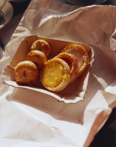 A cardboard tray holds an assortment of small pastries, resting on crumpled white paper. The pastries vary in color and texture, with some topped with sugar crystals and others covered in smooth, layer-like icing. The lighting casts soft shadows, highlighting the pastries’ round shapes and inviting appearance. The scene conveys a cozy, inviting feel perfect for a casual snack.
