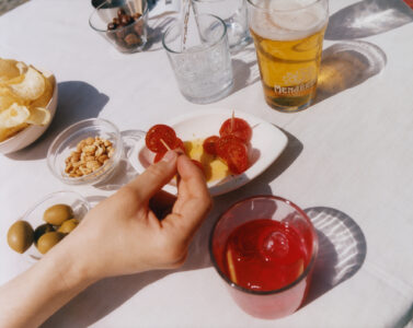 A hand holds sliced tomatoes on a toothpick. Nearby are bowls containing peanuts, olives, and potato chips. Two drinks, one clear and one red with ice and a slice of citrus, sit on a white surface. A glass of amber liquid creates a shadow pattern, suggesting a relaxed outdoor setting with snacks and beverages.