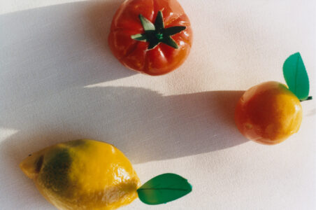 Three glossy, realistic-looking molded fruits rest on a textured surface. The top fruit resembles a red tomato with a detailed green stem, casting a shadow. Below, a yellow lemon with a green leaf cap sits next to an orange with a similar leaf. Each fruit's shiny surface reflects light, adding depth and vibrancy to the arrangement.