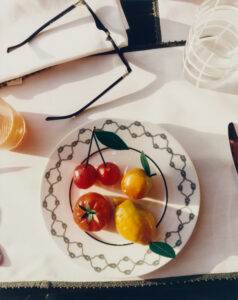 A decorative plate adorned with intricate patterns displays an assortment of brightly colored, lifelike artificial fruits. Nearby, a pair of stylish sunglasses rests on a folded napkin, casting shadows. Beside the plate is a textured glass containing a clear liquid, while warm lighting creates an inviting atmosphere. Various table elements suggest an elegant dining setting.