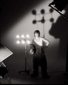 A person stands in a studio, wearing a half-taken-off gorilla suit, revealing a tank top beneath. Bright stage lights are directed at them, casting large shadows on the wall. The scene has a film set atmosphere, with equipment like stands and light panels visible. The black and white composition creates a dramatic and theatrical setting.