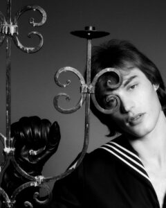 A young person with medium-length hair poses beside an ornate wrought iron structure, contributing to a mysterious and artistic atmosphere. They wear a sailor-style outfit with striped detailing at the collar. One hand, gloved in black, rests against the ironwork, contrasting with the smooth backdrop. The lighting casts soft shadows, enhancing the monochromatic and stylish essence.