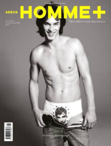 A person with dark hair stands confidently, smiling directly. They are topless, wearing a pair of distinctive underwear adorned with a face and star design. Jeans are loosely held around the hips. The background and overall style convey a sense of bold fashion and contemporary style, capturing a playful and edgy aesthetic.