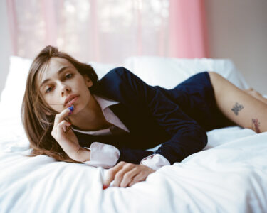 A person with long brown hair reclines on a white bed, wearing a dark blazer over a white shirt. Their gaze is directed at the viewer as they rest their chin on one hand, displaying nails painted in a vibrant color. Behind them, soft pink curtains diffuse light, creating a relaxed, intimate atmosphere. Tattoos are visible on their upper hip.