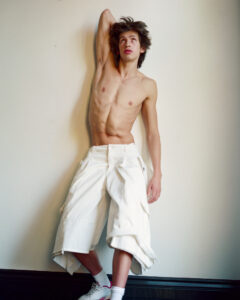 An individual stands against a plain wall, leaning slightly backwards. With one arm raised behind their head, they gaze upwards contemplatively. They have a lean, toned physique and wear baggy, knee-length white shorts with large pockets. Their sneakers are white with hints of pink, complemented by white socks. The ambient lighting is soft, creating subtle shadows.