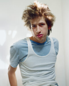 A young person with tousled hair exudes a casual, slightly rebellious vibe. They wear a striped blue t-shirt layered with a white sleeveless top. The gaze is direct, conveying a mix of confidence and contemplation. Soft natural light enhances the relaxed atmosphere, casting gentle shadows and highlights across the face and background, creating a fresh, candid feel.