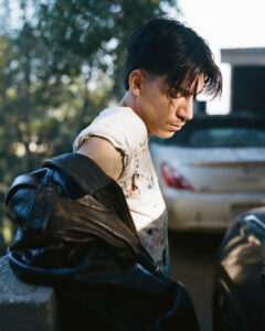 A young man with dark hair looks thoughtful, partially wearing a leather jacket over a paint-splattered shirt. He stands outdoors, surrounded by blurred greenery and parked cars under bright sunlight. His expression is pensive, as light and shadows play across his features. The scene conveys a casual, contemplative moment captured in a serene atmosphere.