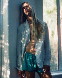 A person with long hair leans against a wall, bathed in dappled sunlight filtering through a nearby window. They wear a loose, white jacket over their shoulders and teal shorts. The atmosphere is serene, with soft shadows creating intricate patterns on their skin. The window reveals glimpses of greenery outside, enhancing the tranquil, natural vibe.