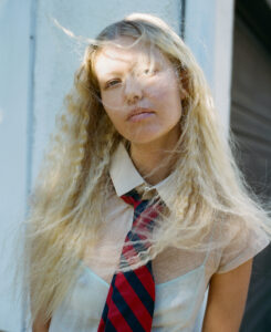 A person with long, wavy blonde hair stands in semi-shade, sunlight creating delicate patterns on their face. Their expression is calm and contemplative. They wear a light-colored short-sleeved blouse and a vibrant red and blue striped tie. The setting includes a bright wall and a shadowy area, suggesting a mix of indoor and outdoor elements. The overall mood is serene and artistic.