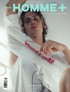 A young person with tousled hair gazes downward while lifting a loose white shirt over one shoulder, revealing part of the torso and underarm. Soft natural light washes across the scene, creating a dreamy, intimate atmosphere. Relaxed body language and muted tones convey effortless confidence, vulnerability, and quiet sensuality, blending casual elegance with youthful energy and charm.