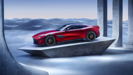 A sleek red sports car is prominently displayed on a modern, angular platform. The vehicle's polished bodywork gleams against the minimalist backdrop of muted blue and grey tones. The scene is set in a surreal, dreamlike environment with distant misty hills and vertical architectural elements framing the composition, enhancing the car's elegance and dynamic presence.