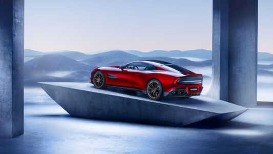 A sleek, red sports car with a streamlined design is positioned on an angular platform, set against a backdrop of rolling, misty blue hills. The car's smooth curves and shiny exterior exude luxury and speed. The setting provides a futuristic, elegant ambiance, highlighted by the contrast between the vibrant car and the serene landscape.