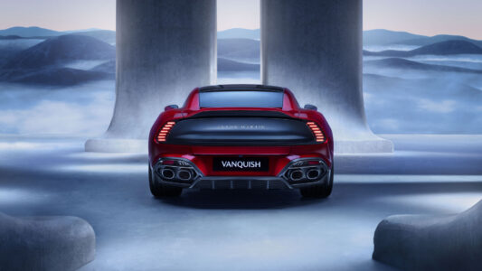 A sleek, red sports car is parked in an elegant, futuristic courtyard. The vehicle's rear view showcases its aerodynamic design with prominent taillights and dual exhausts. The background features massive concrete pillars and a serene, misty landscape with undulating hills. The setting combines modern architectural elements with a tranquil, blue-tinted atmosphere, highlighting the car's sophisticated appeal.