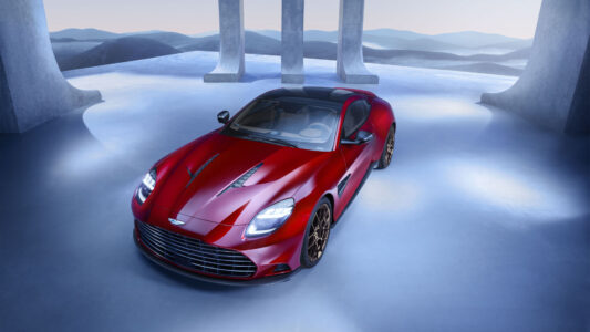 A sleek, red sports car is positioned under an open structure with tall, abstract pillars. The car's glossy finish reflects the soft, ambient lighting, highlighting its aerodynamic design and polished, bronze wheels. The background features a serene, undulating landscape in shades of blue and gray, creating a contrast that emphasizes the vehicle's bold color and dynamic form.