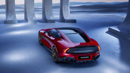 A sleek red sports car is parked on a glossy, reflective surface, surrounded by tall, grey stone pillars. The setting feels futuristic and serene, with a soft blue hue dominating the background, suggesting a distant horizon. The car, with its aerodynamic design and dark sunroof, is positioned slightly to the left, highlighting its elegance and modernity.