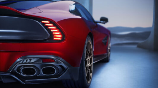 A sleek, red sports car is showcased from the rear, highlighting its aerodynamic design. The vehicle boasts dual exhausts and distinctive vertical taillights. The wheels have an intricate gold alloy design, emphasizing luxury and performance. The setting appears minimalistic, with soft lighting creating a contrast against the car's glossy finish, suggesting modernity and elegance.