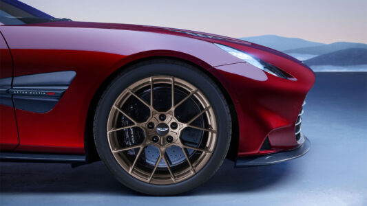 A sleek, red sports car exudes elegance with its smooth curves and glossy finish. The focus is on its front section, where an intricate, gold-colored alloy wheel stands out. The background features a serene, muted landscape with soft, gradient hues of blue and gray, hinting at distant hills under a calm sky, creating a dynamic yet tranquil scene.