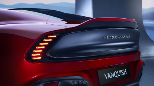A sleek, red sports car showcases its dynamic rear design, highlighted by distinctive, illuminated tail lights that form a modern, horizontal pattern. The aerodynamic shape is emphasized by a subtle spoiler, adding a sense of speed and elegance. The background features blurred, cool-toned elements, suggesting a setting that contrasts with the car's vibrant color and polished finish.