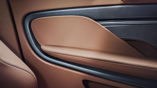 The interior detail showcases a sleek car door panel featuring a combination of smooth brown leather and textured carbon fiber accents. The design includes elegant curves and edges, with a subtle indentation running horizontally across the center. The contrast between the warm leather tones and the dark, glossy carbon fiber evokes a luxurious, modern aesthetic.